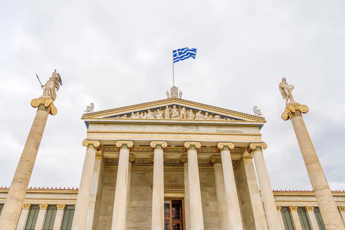Die Nationale Akademie von Athen steht im Tageslicht unter einem strahlend blauen Himmel; vom Dach weht stolz eine griechische Flagge