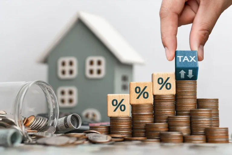 Um das Konzept der Grundsteuer zu veranschaulichen, legt eine Hand einen Würfel mit der Aufschrift „TAX“ auf einen Stapel Münzen in der Nähe eines Modellhauses.