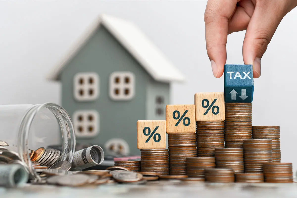 Um das Konzept der Grundsteuer zu veranschaulichen, legt eine Hand einen Würfel mit der Aufschrift „TAX“ auf einen Stapel Münzen in der Nähe eines Modellhauses.